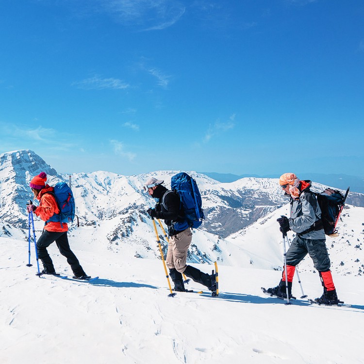Three hikers using 4-in-1 lightweight terrain snowshoes on snowy mountains, equipped with ski poles and backpacks.